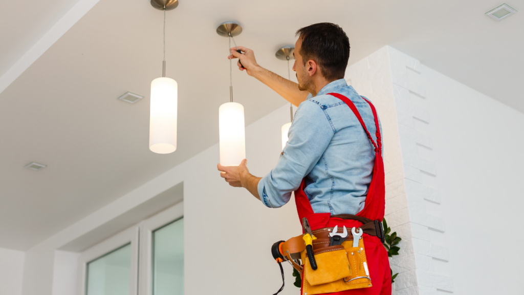 A man in red overalls and a toolbelt is installing clean white light fixtures on a ceiling.